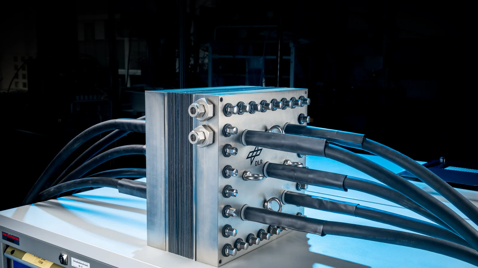 Electrolyser short stack with a bolted metal end plate, lifting eyes and insulated hoses, dramatically lit in a test hall.