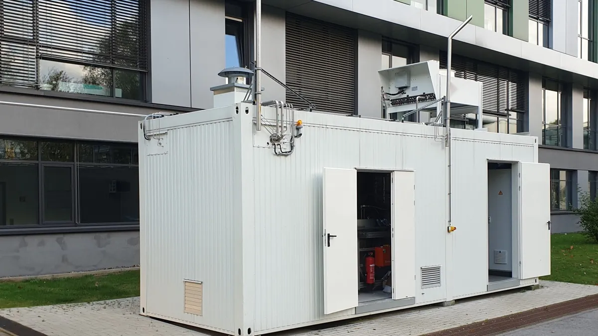 Containerised water electrolyser unit installed outdoors next to a building, with vent stack and cooler on the roof